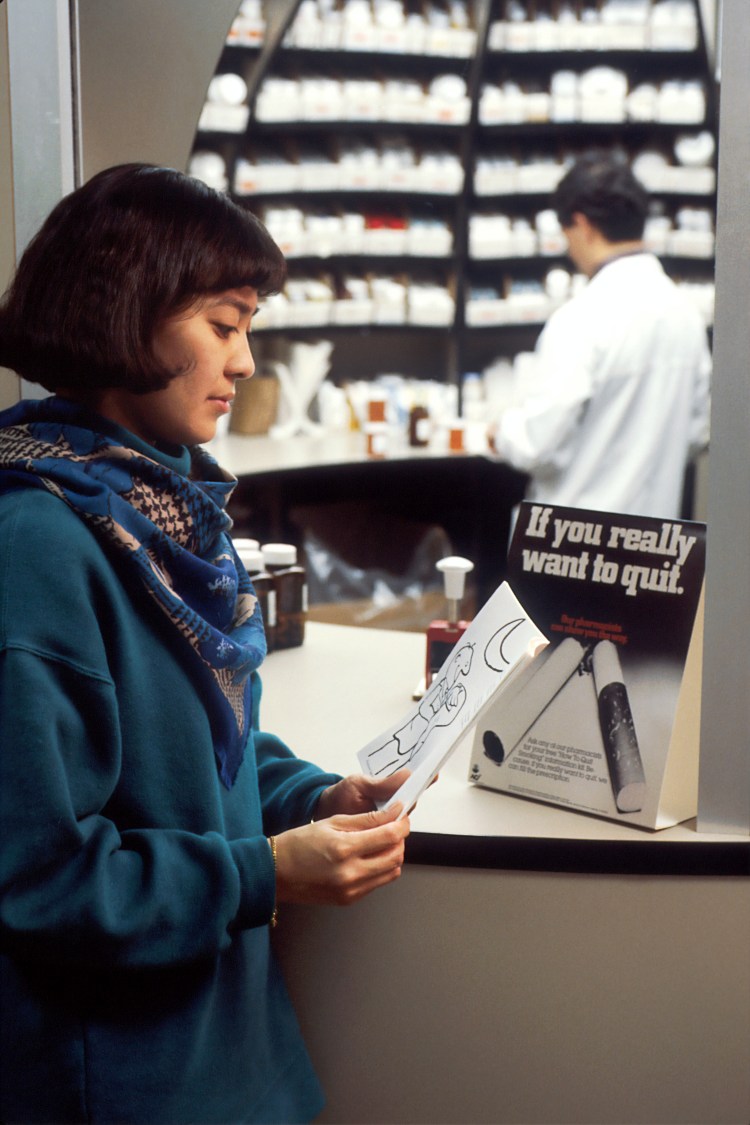 An Asian female reading a booklet at a pharmacy counter while a male pharmacist works in the background.